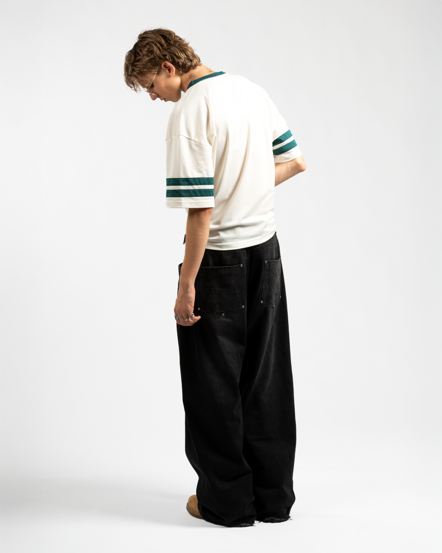 Back view of Whillie Clothing’s men’s black baggy jeans featuring tonal swirl embroidery on the rear pockets and an ultra-relaxed wide-leg fit, paired with a vintage mesh jersey and tan boots