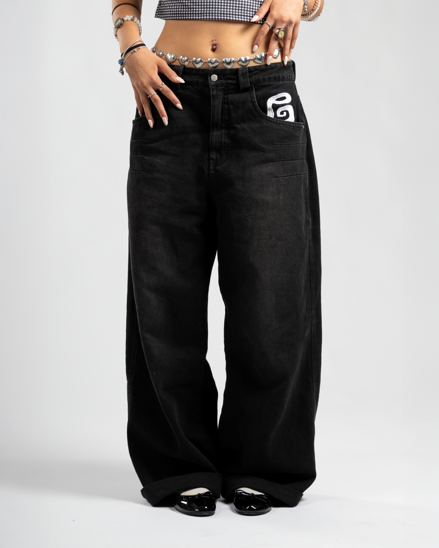Front view of Whillie Clothing’s women’s black baggy jeans featuring a bold white embroidered logo on the pocket, styled with a heart chain belt and glossy black flats for a Y2K-inspired streetwear vibe