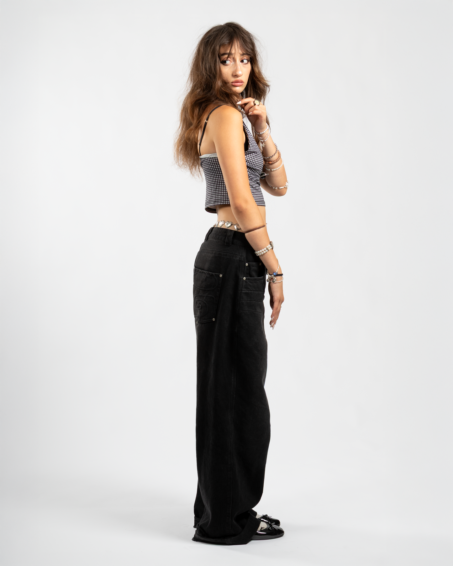 Side view of Whillie Clothing’s women’s black baggy jeans showcasing tonal swirl embroidery on the back pocket, styled with a cropped checkered top and ballet flats