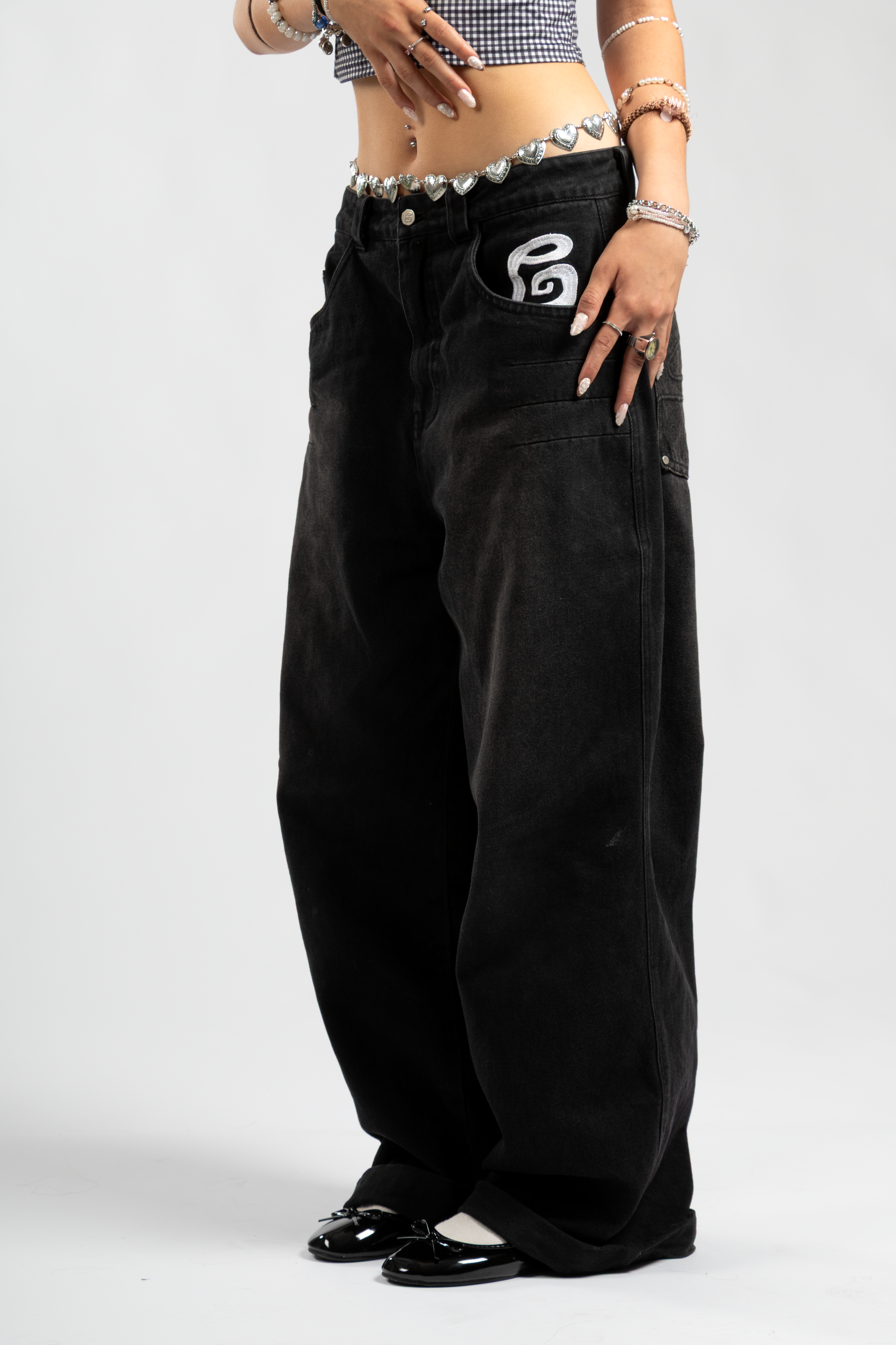 Front view of black wide-leg baggy jeans with bold white embroidered pocket logo, styled with a silver heart chain belt and glossy black ballet flats