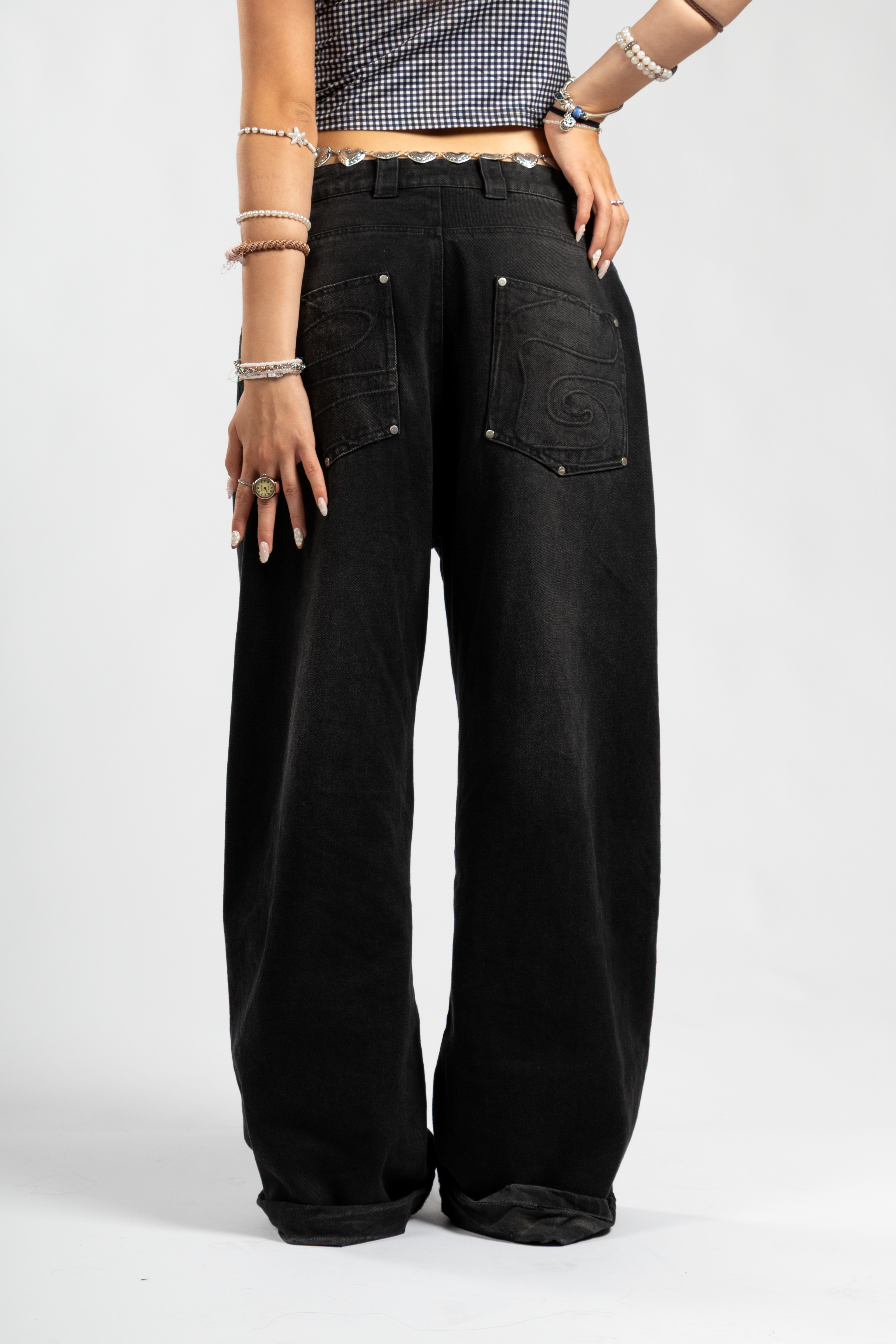 Back view of black baggy jeans with oversized fit and tonal embroidered swirl back pockets, styled with a silver heart chain belt and checkered crop top