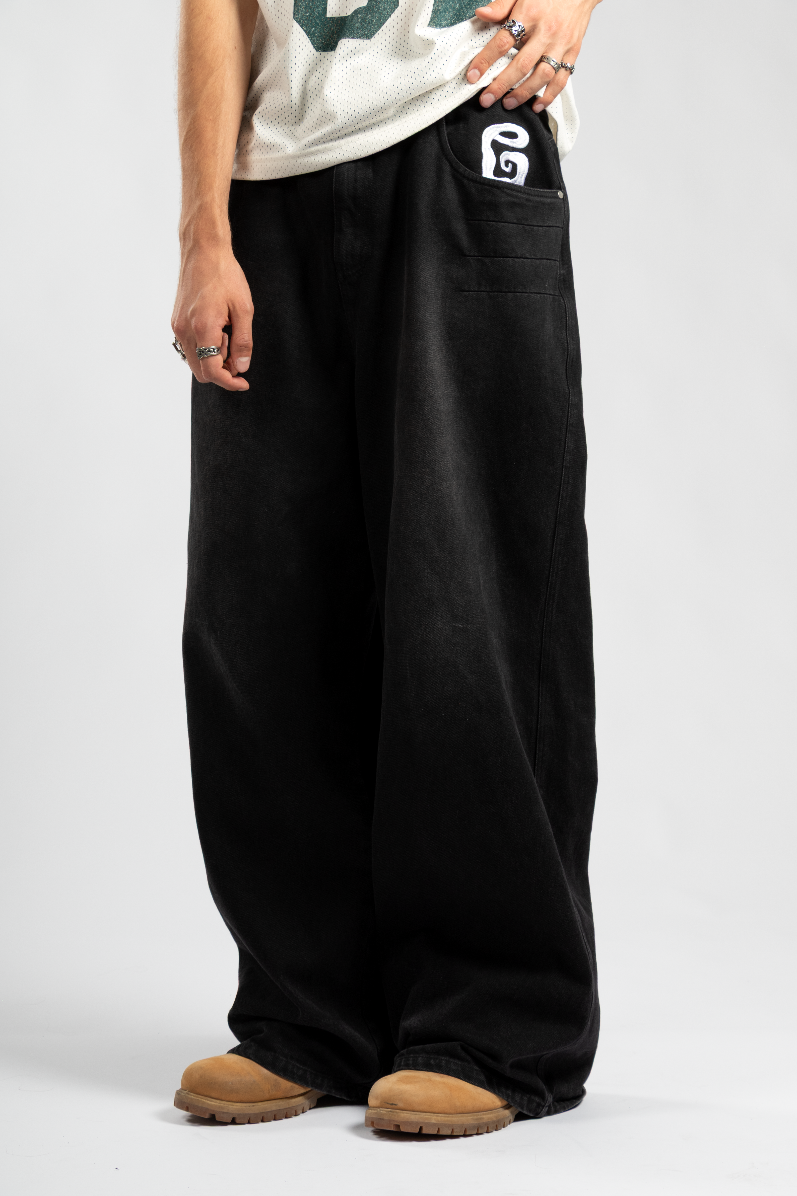 Front view of men's black baggy jeans with white embroidered pocket detail, paired with a mesh jersey tee and tan work boots for a relaxed streetwear look