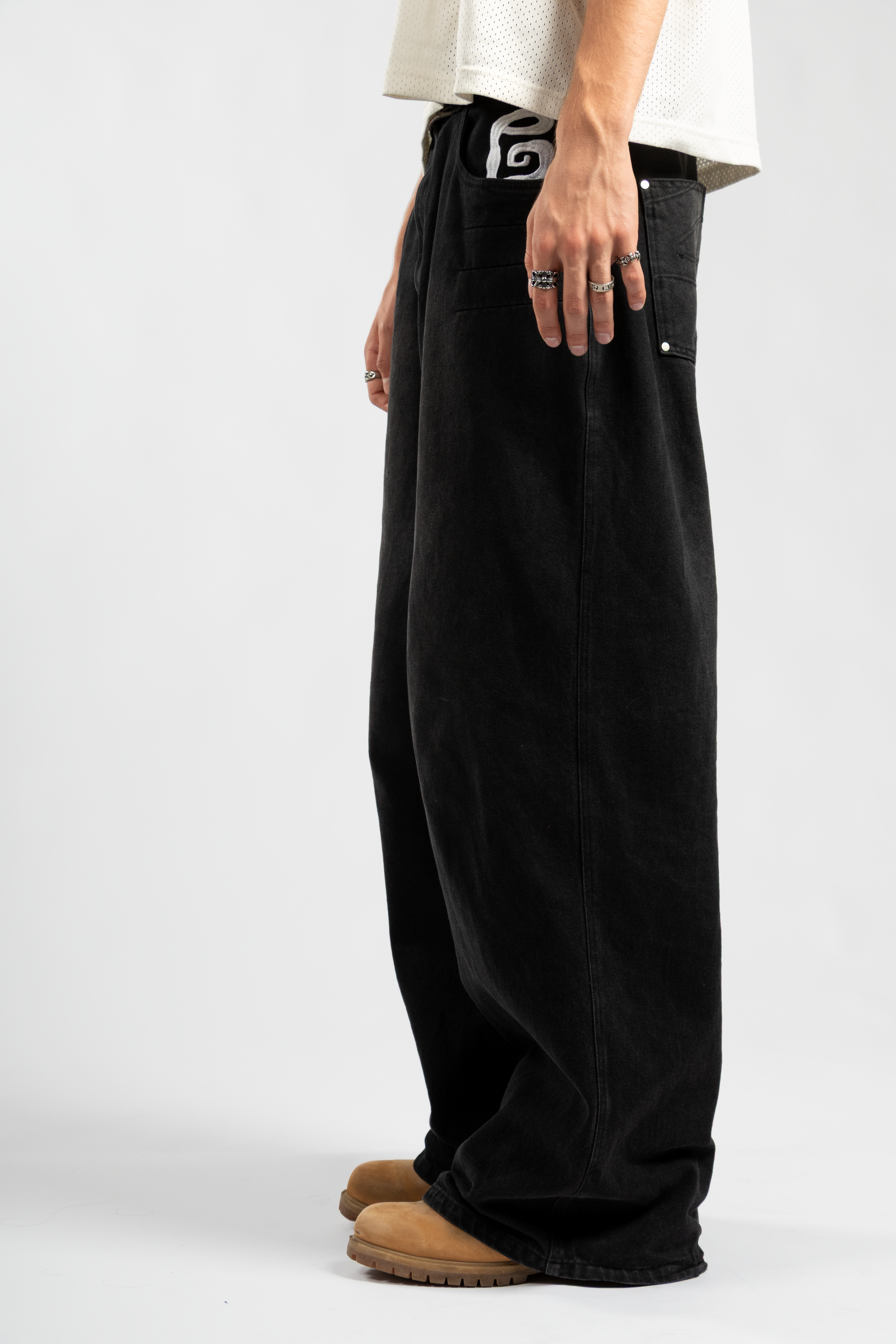 Side view of black baggy jeans with white embroidered graphic peeking from front pocket, styled with tan work boots and silver rings for a rugged streetwear aesthetic