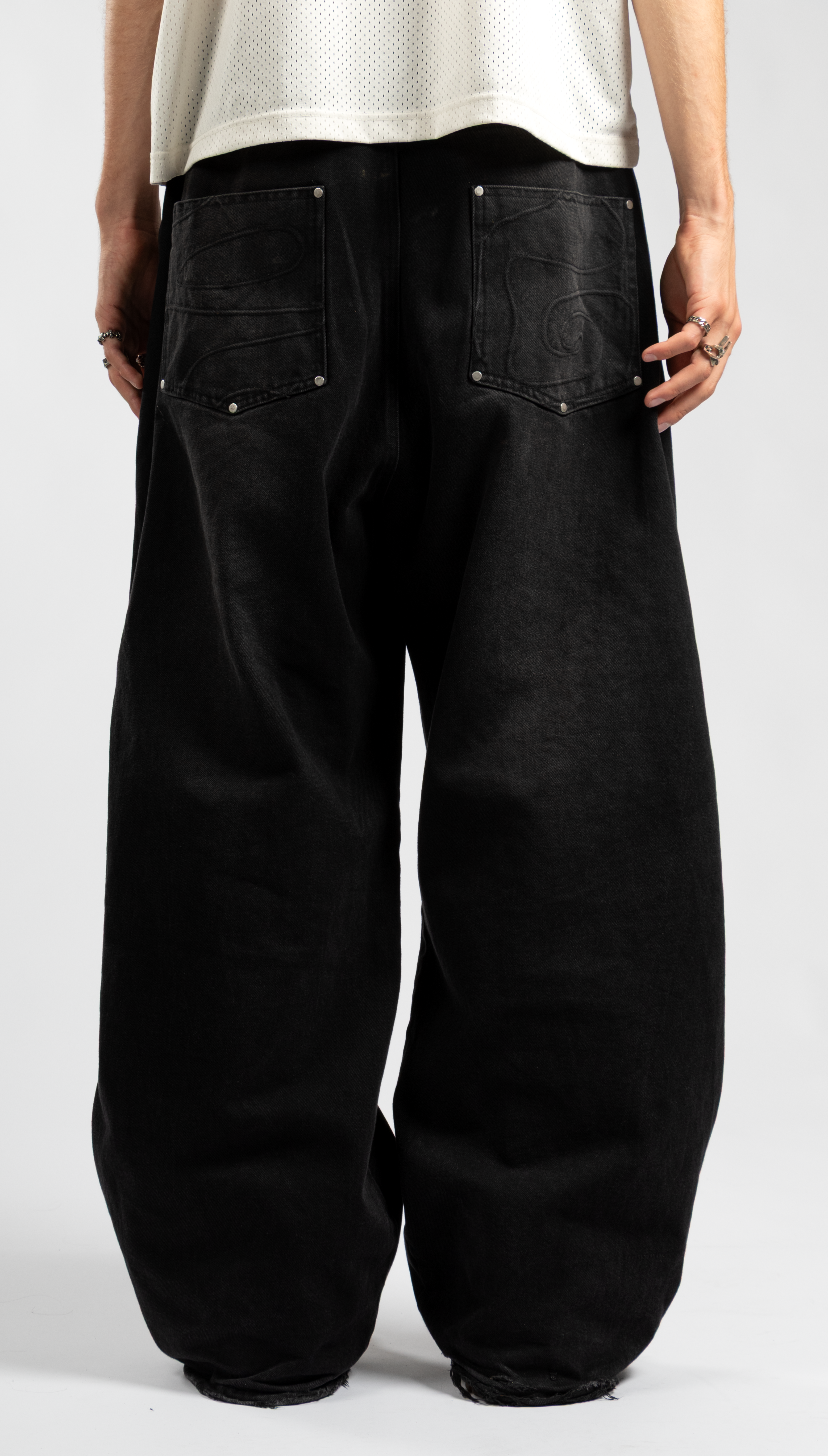 Behind view of men's oversized black baggy jeans featuring tonal embroidered swirl back pockets and extra-wide leg fit, styled with a mesh jersey tee on a white background