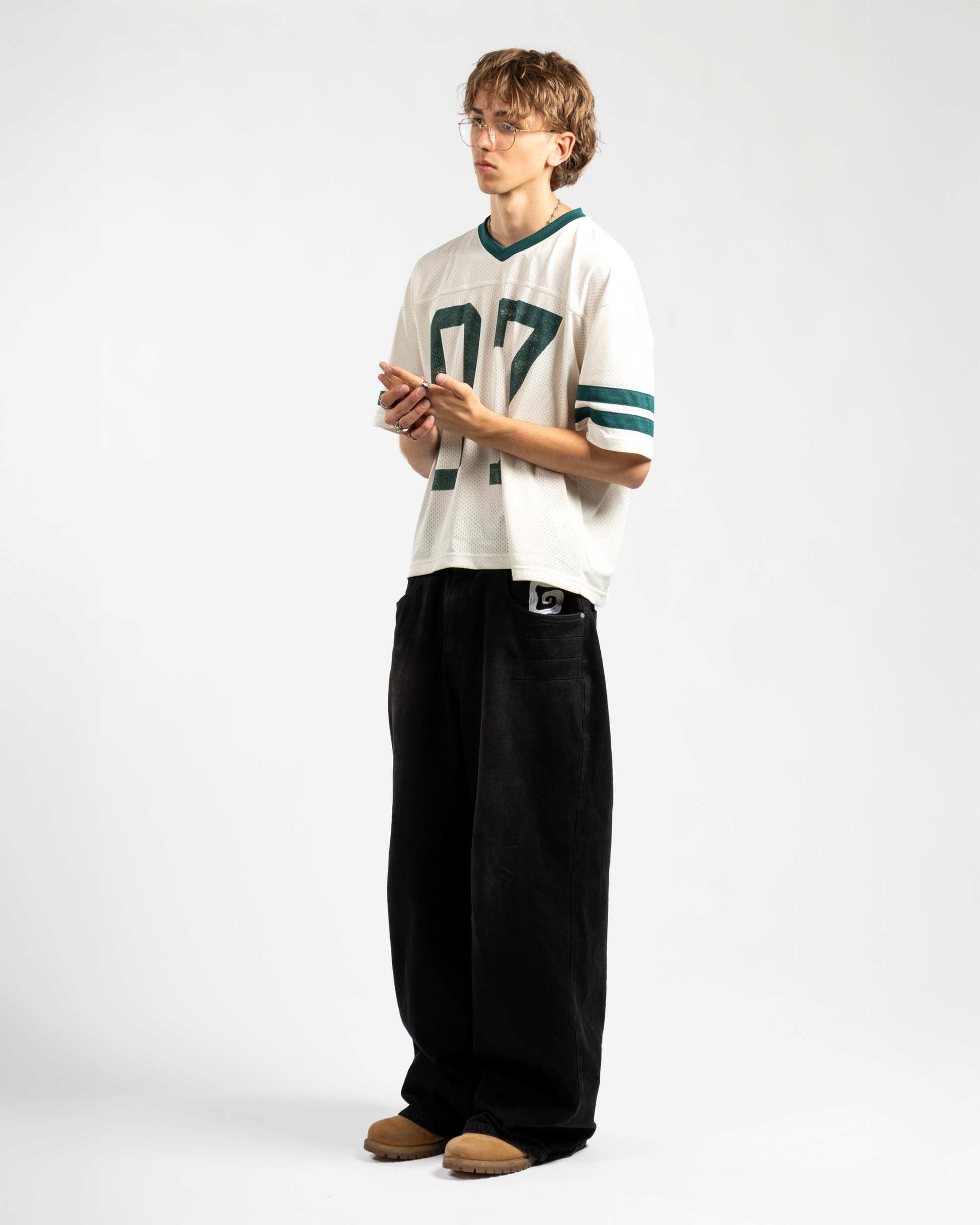 Full body view of Whillie Clothing’s men’s oversized black baggy jeans featuring a bold white embroidered logo on the front pocket, styled with a mesh football jersey and tan boots for a modern retro streetwear fit