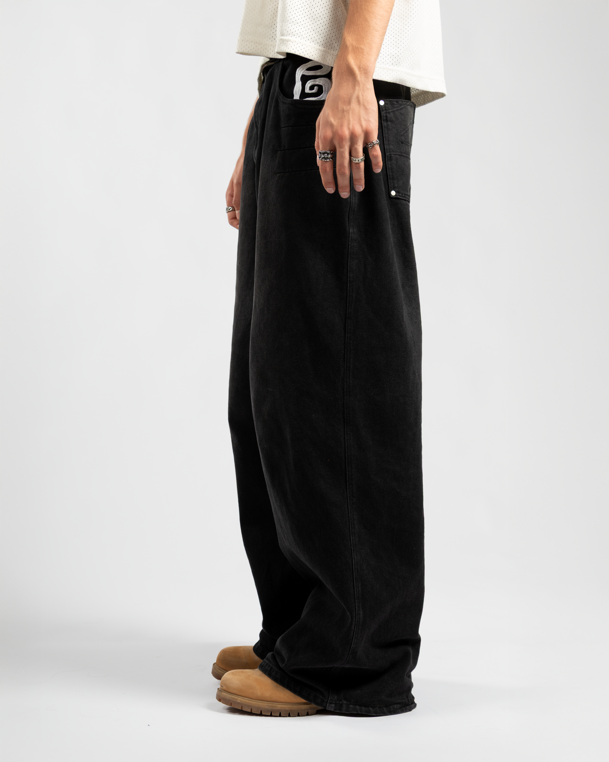 Side view of Whillie Clothing’s oversized black baggy jeans for men, featuring a deep front pocket and signature white embroidered logo, paired with tan boots for a rugged streetwear look