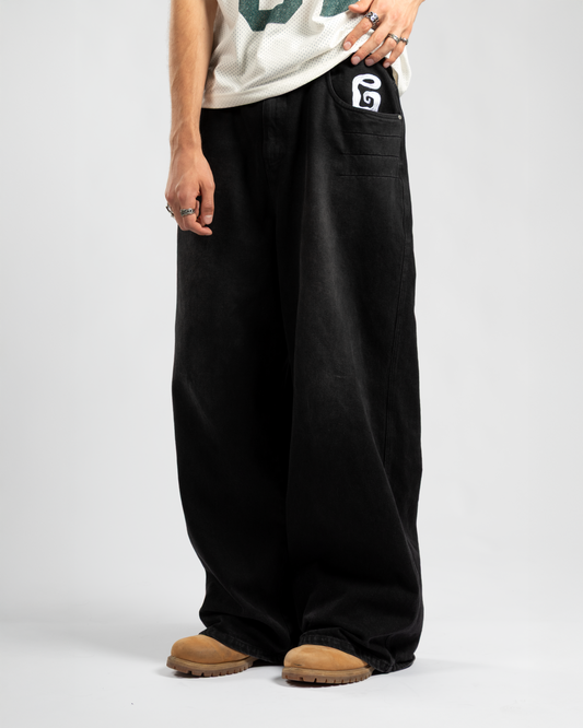 Front view of Whillie Clothing’s signature black baggy jeans with oversized fit and bold white embroidered pocket logo, styled with tan boots and a mesh jersey for a clean streetwear statement