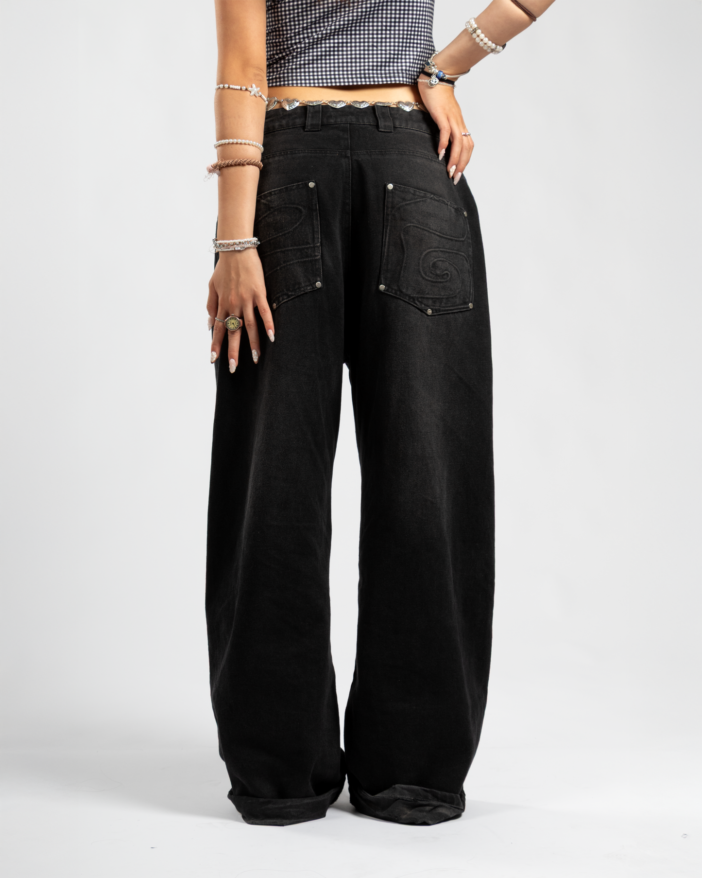 Back view of Whillie Clothing’s women’s black baggy jeans featuring tonal embroidered swirl pockets and an oversized fit, styled with a silver heart belt and checkered crop top