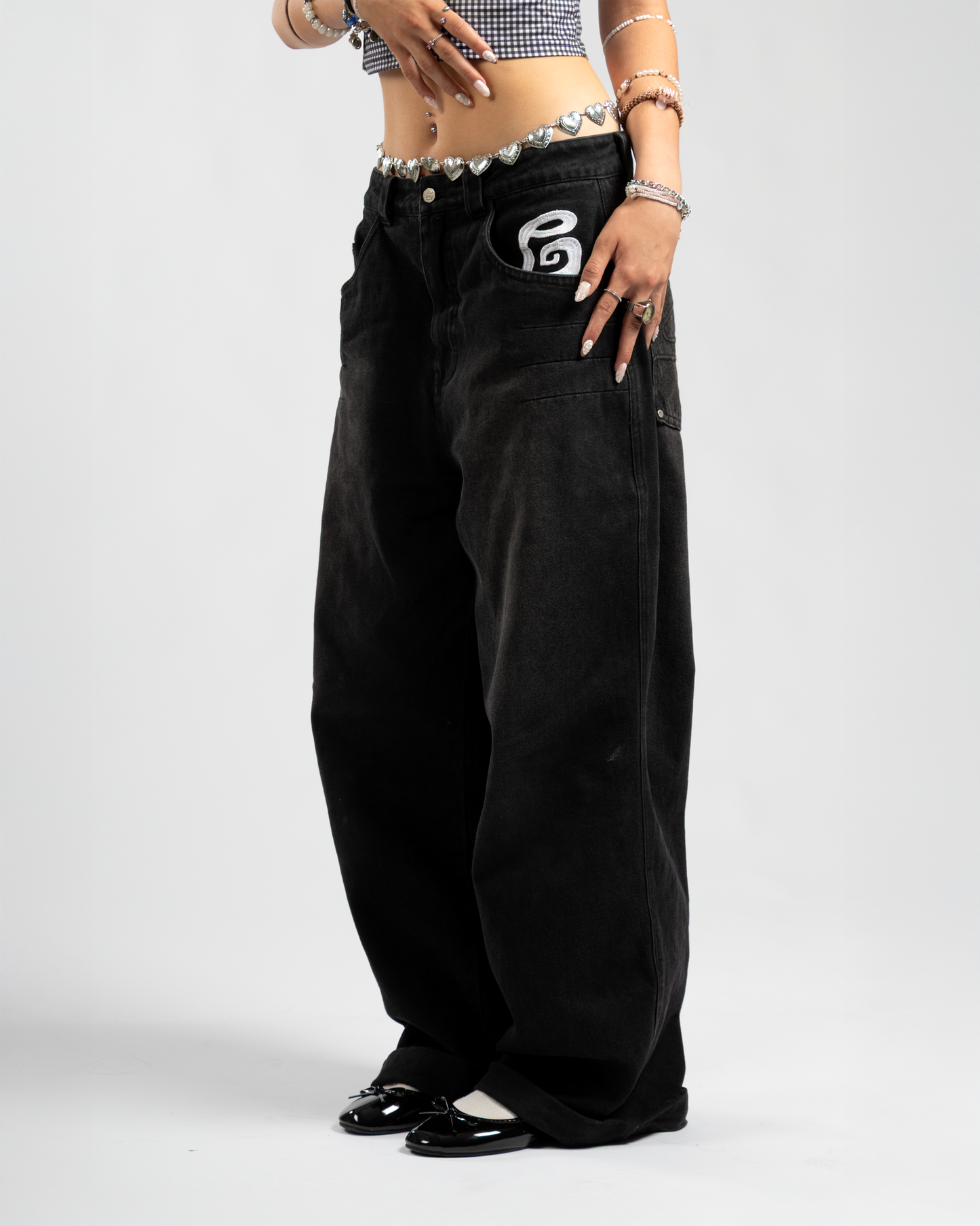 Side view of Whillie Clothing’s women's oversized black baggy jeans with front pocket embroidery, styled with a silver heart chain belt and glossy ballet flats for a bold Y2K streetwear look