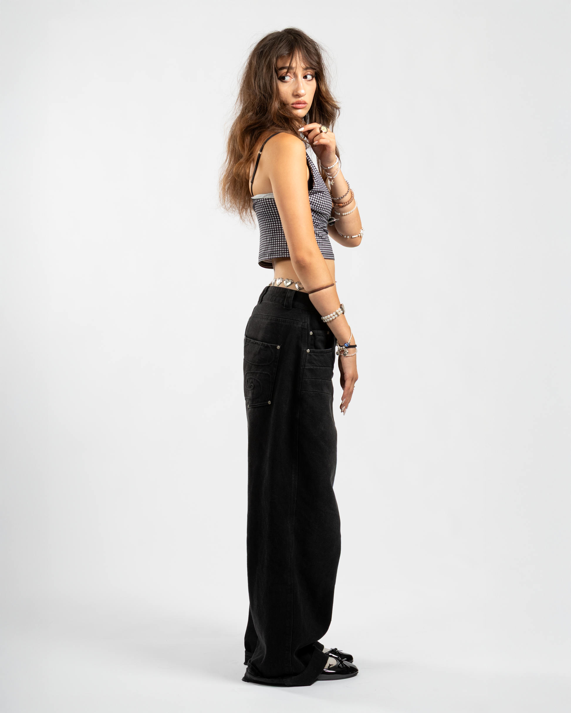 Side view of Whillie Clothing’s women’s black baggy jeans showcasing tonal swirl embroidery on the back pocket, styled with a cropped checkered top and ballet flats