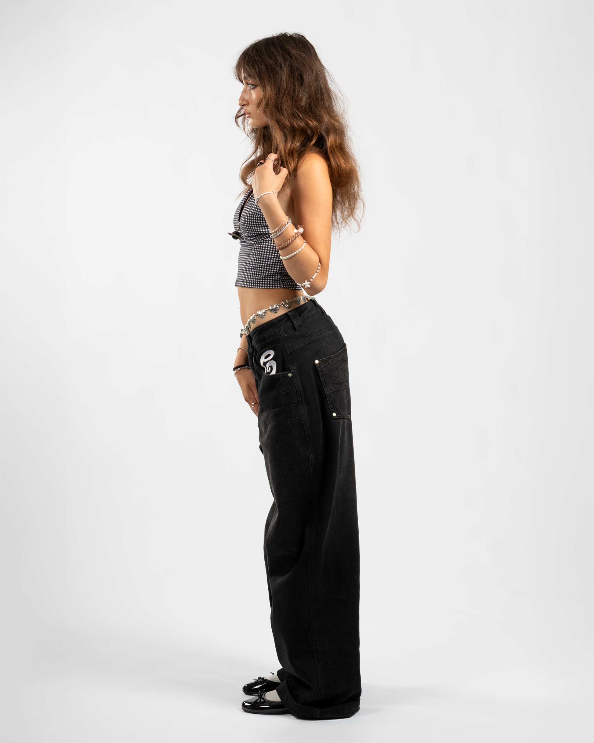 Side view of Whillie Clothing’s women’s black baggy jeans with oversized fit, white embroidered logo on the pocket, and tonal swirl detail on the back pocket — styled with ballet flats and a checkered crop top for a feminine streetwear twist