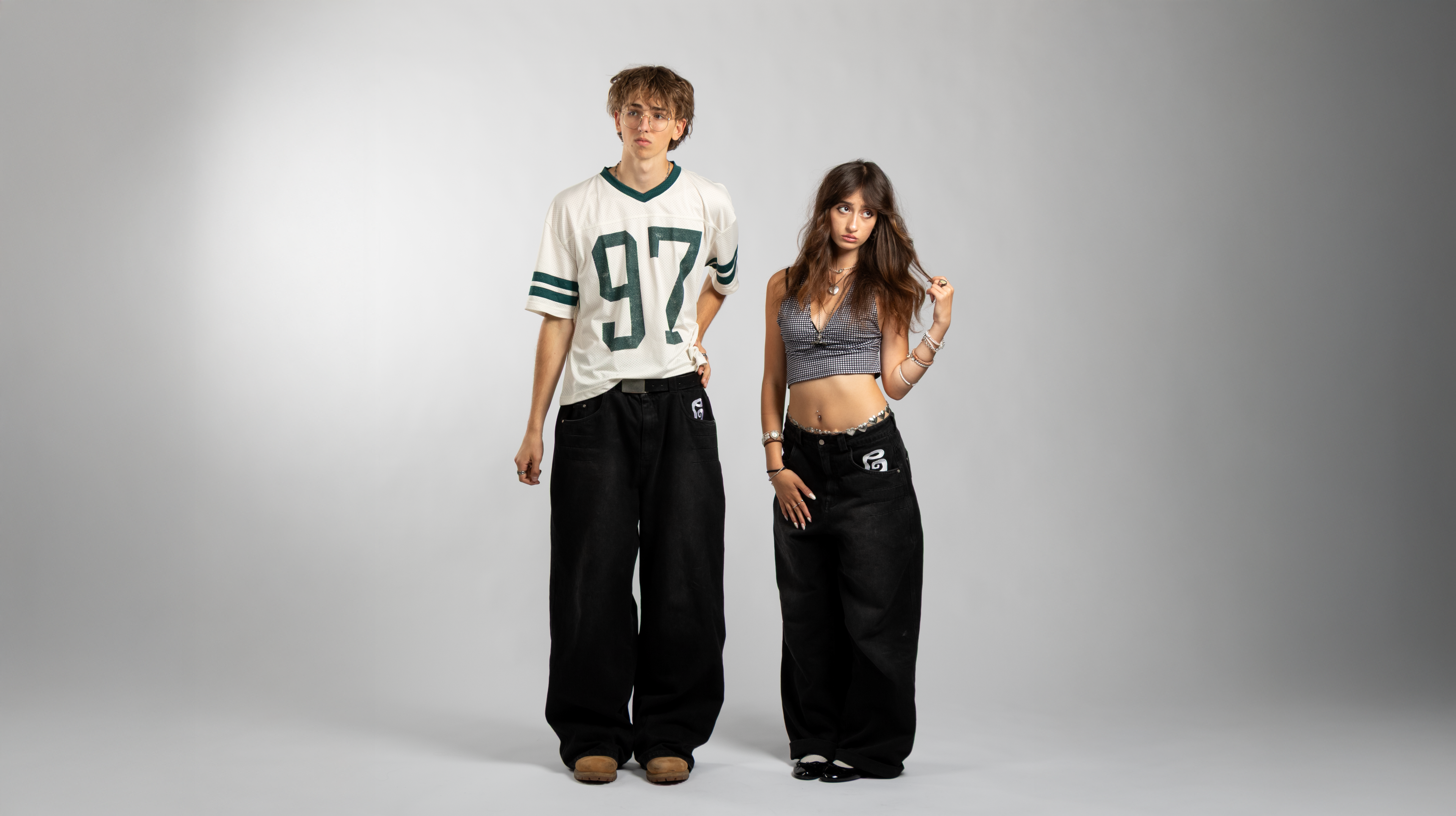 Two models wearing matching oversized black baggy jeans with embroidered front pocket details, highlighting the unisex fit, wide-leg cut, and bold streetwear silhouette — styled with a crop top and mesh football jersey