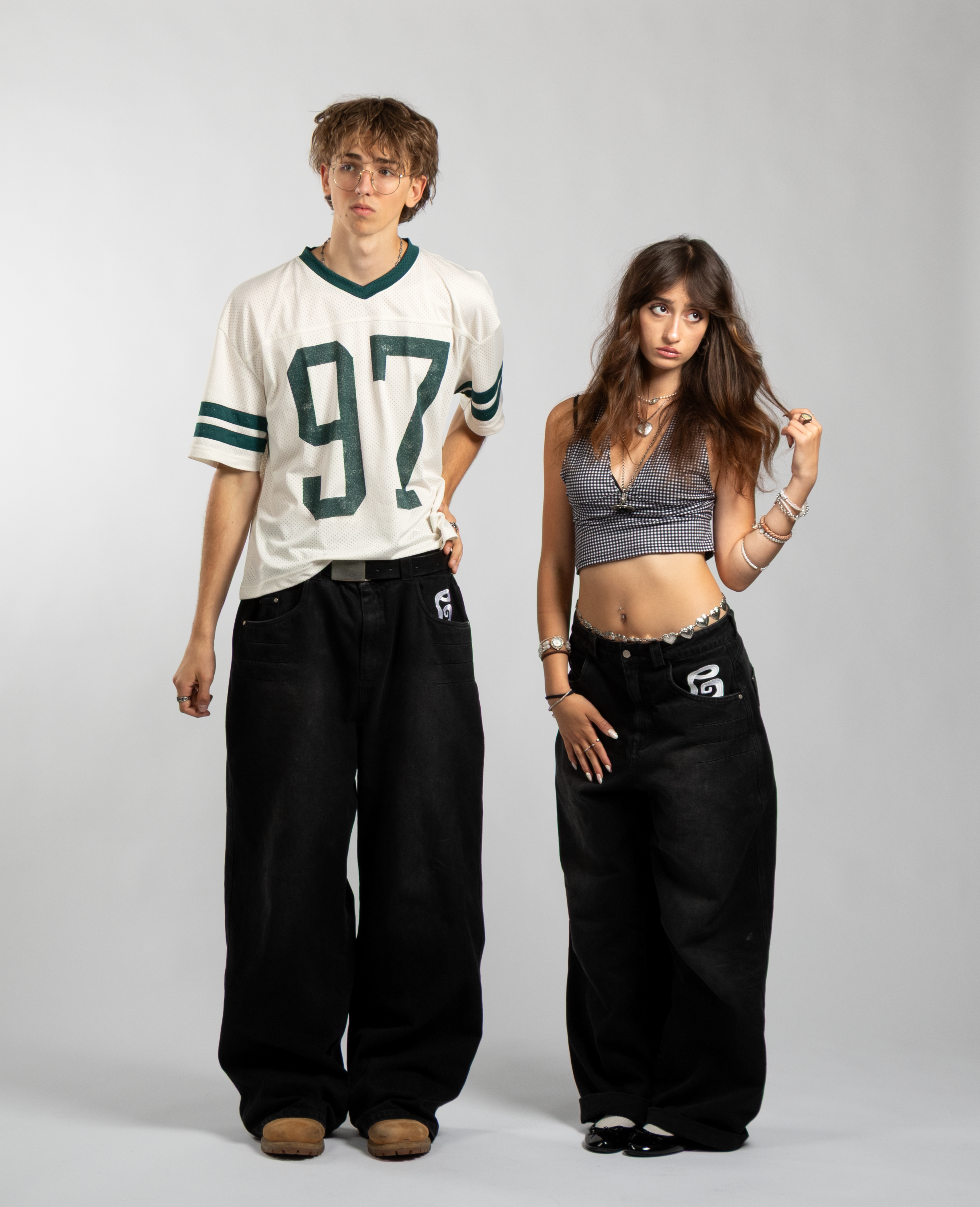 Two models wearing matching oversized black baggy jeans with embroidered front pocket details, highlighting the unisex fit, wide-leg cut, and bold streetwear silhouette — styled with a crop top and mesh football jersey