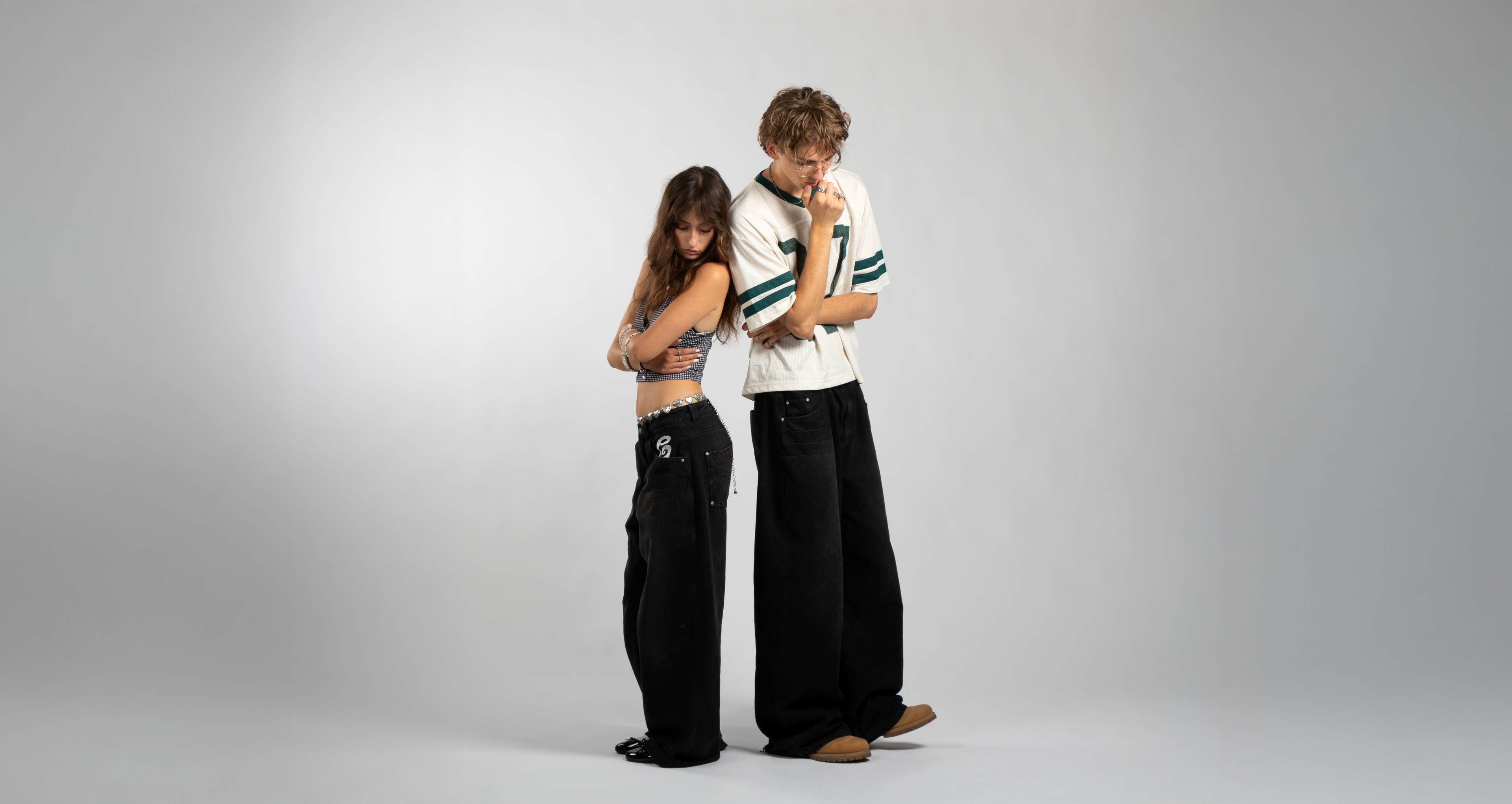 Two models showcasing oversized black baggy jeans with embroidered pocket details, emphasizing the unisex fit and bold streetwear silhouette — styled with a checkered crop top and silver heart belt, and a vintage mesh jersey tee