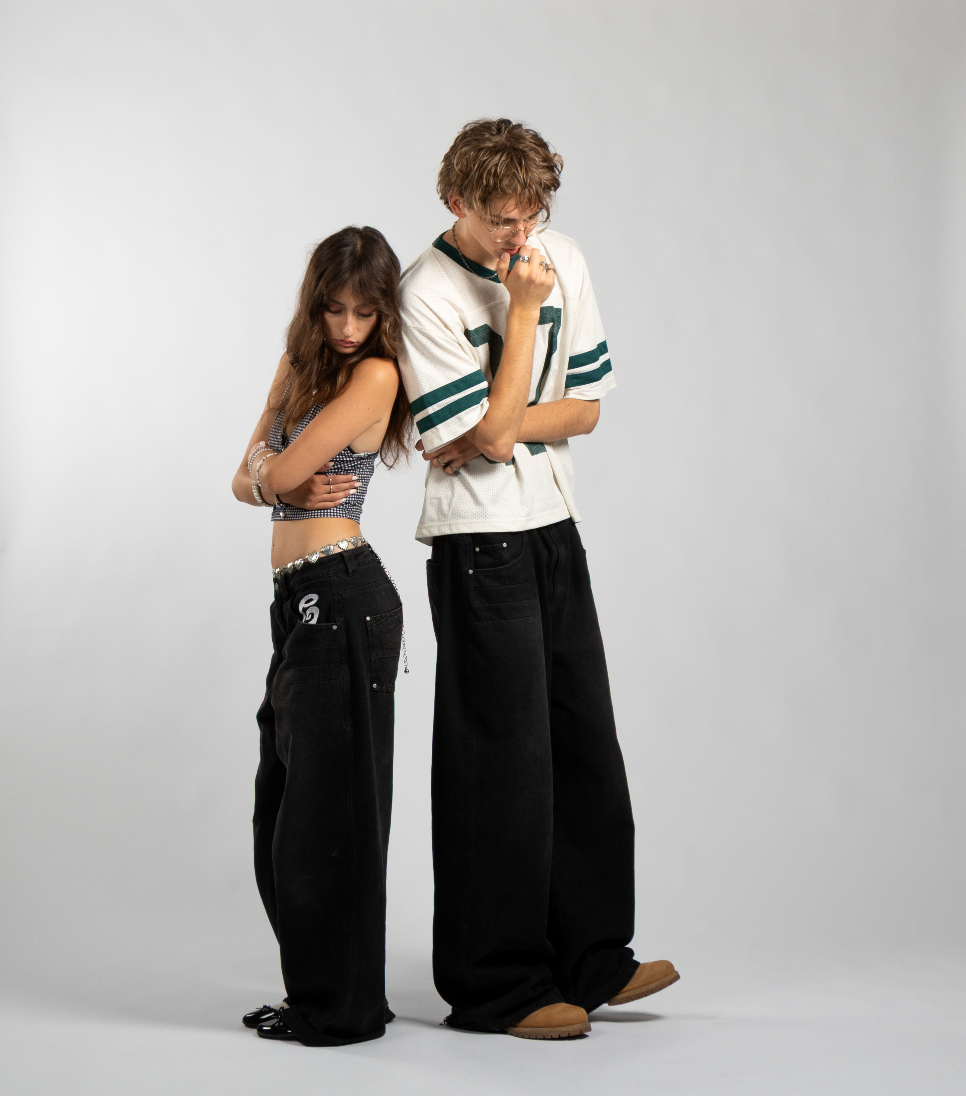 Two models showcasing oversized black baggy jeans with embroidered pocket details, emphasizing the unisex fit and bold streetwear silhouette — styled with a checkered crop top and silver heart belt, and a vintage mesh jersey tee
